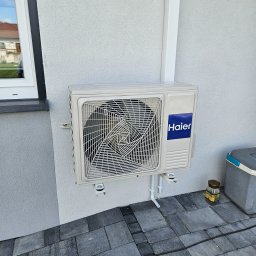 Novatech Energy sp. z o.o. - Zewnętrzna jednostka klimatyzacji Haier zamontowana na elewacji domu, widoczne rurki odprowadzające skropliny i puszka po kawie Jacobs na kostce brukowej.