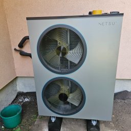 Novatech Energy sp. z o.o. - Szara pompa ciepła NETSU z dwoma wentylatorami, zamontowana na zewnątrz budynku, widoczny fragment elewacji i izolowane rury, na górze leży taśma i narzędzie wielofunkcyjne, obok stoi zielone wiadro.
