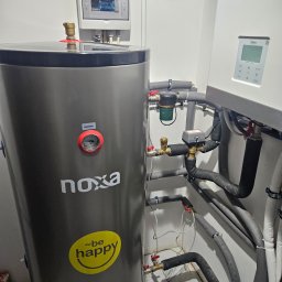 Novatech Energy sp. z o.o. - Szary zasobnik ciepłej wody użytkowej marki Noxa z naklejką 'Be Happy', podłączony do instalacji hydraulicznej z licznymi rurami, zaworami i filtrem, obok wisząca pompa ciepła Midea i panel...