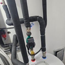Novatech Energy sp. z o.o. - Instalacja grzewcza: rury w czarnej izolacji, zawory z kolorowymi oznaczeniami, pompa obiegowa Wilo zamontowana na białym zbiorniku, widoczne elementy instalacji hydraulicznej.