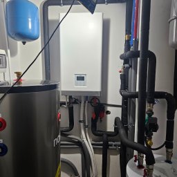 Novatech Energy sp. z o.o. - Węzeł cieplny z kotłem gazowym, zbiornikiem buforowym i zasobnikiem ciepłej wody użytkowej, rury w izolacji termicznej, manometr na niebieskim zbiorniku wyrównawczym.