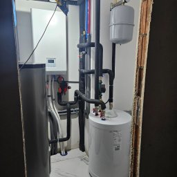 Novatech Energy sp. z o.o. - Węzeł cieplny w pomieszczeniu z wykończonymi ścianami, widoczne rury w izolacji, zbiorniki, oraz biała jednostka sterująca na ścianie, oświetlenie robocze zawieszone na rurze.