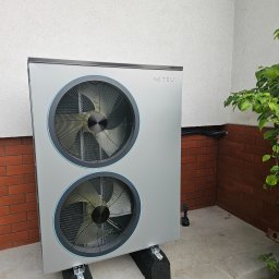 Novatech Energy sp. z o.o. - Zewnętrzna jednostka pompy ciepła NETSU z dwoma wentylatorami, zamontowana na czarnych podkładkach, z widocznymi przewodami na tle białej i ceglanej ściany budynku.