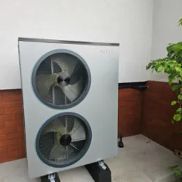 Zewnętrzna jednostka pompy ciepła NETSU z dwoma wentylatorami, zamontowana na czarnych podkładkach, z widocznymi przewodami na tle białej i ceglanej ściany budynku.