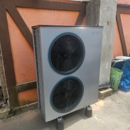 Novatech Energy sp. z o.o. - Zewnętrzna jednostka pompy ciepła NIBE ustawiona na gumowych nóżkach, widoczna na tle pomarańczowej ściany budynku z elementami dekoracyjnymi w kolorze brązowym.