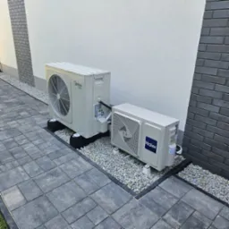 Dwie jednostki zewnętrzne klimatyzacji, Midea i Haier, ustawione na podłożu z szarego żwiru przy ścianie budynku z elementami dekoracyjnymi z cegły.