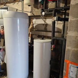 Novatech Energy sp. z o.o. - Dwa białe zasobniki na wodę o różnych rozmiarach w pomieszczeniu z betonowymi ścianami i widocznymi instalacjami hydraulicznymi, w tle fragment sylwetki osoby i psa.