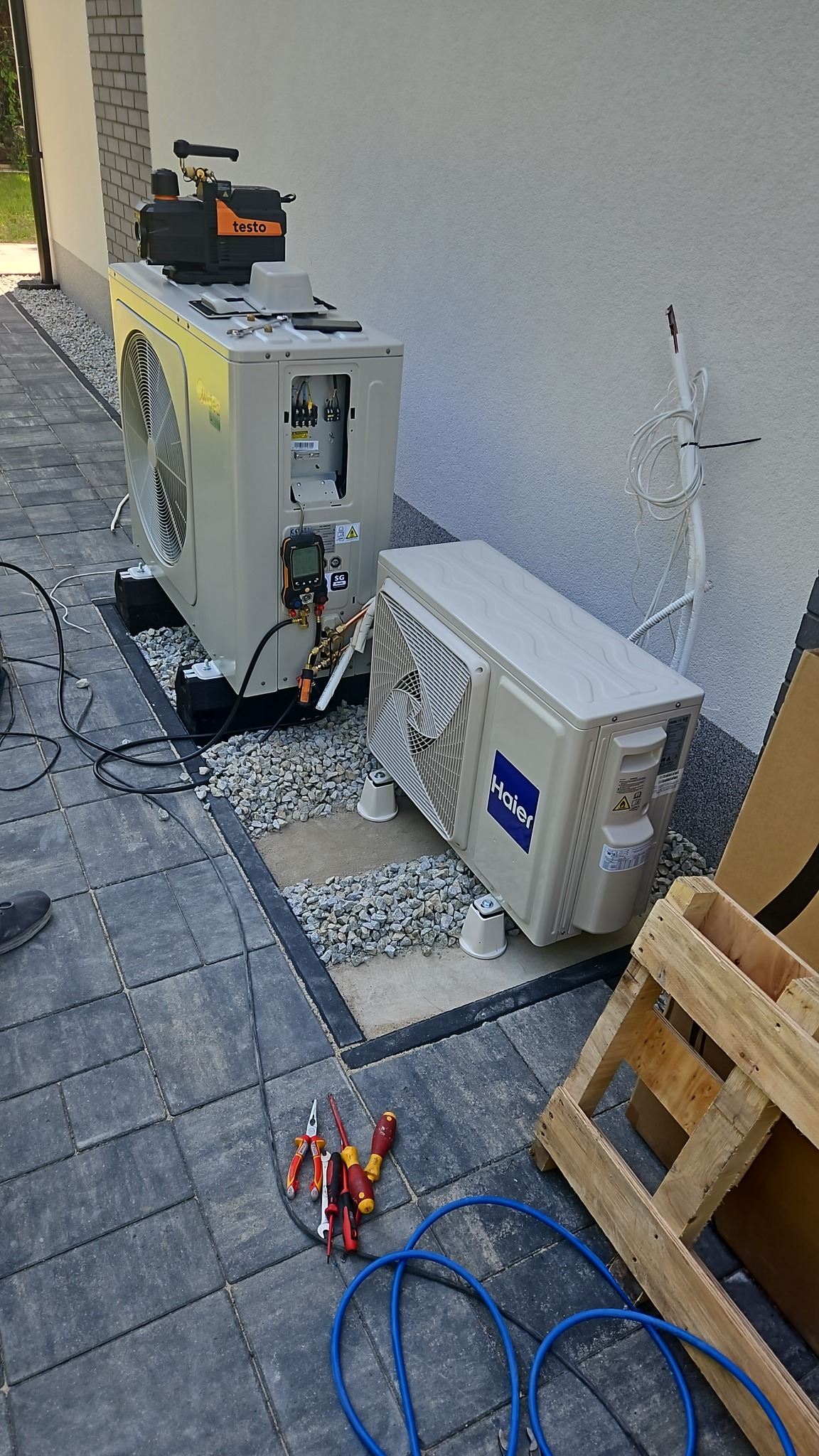 Dwie jednostki zewnętrzne klimatyzacji, Midea i Haier, ustawione na żwirowym podłożu przy białej ścianie budynku, z narzędziami (szczypce, śrubokręty) i niebieskimi przewodami leżącymi na szarej...