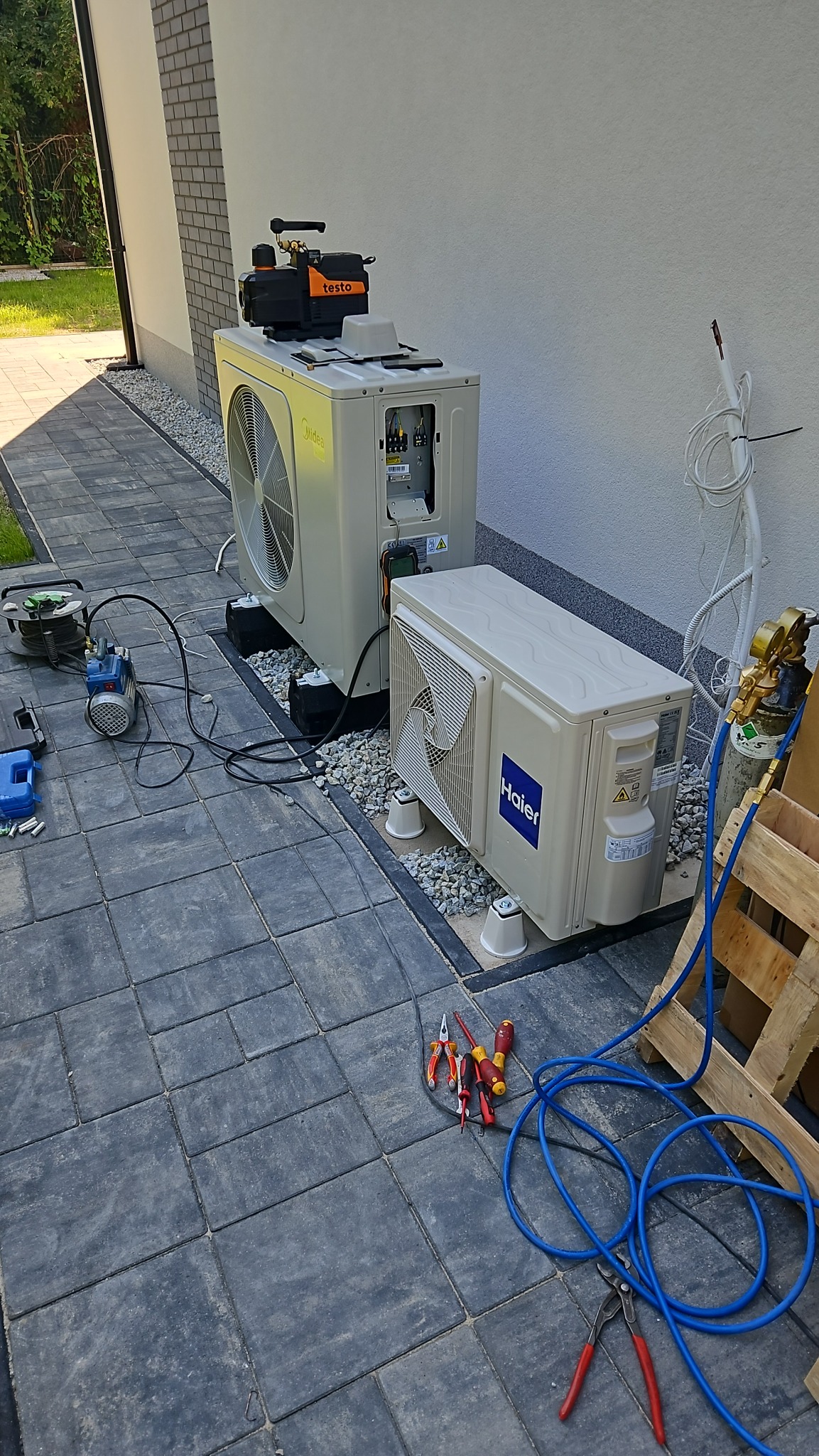 Dwie jednostki zewnętrzne klimatyzacji, jedna marki Midea, druga Haier, połączone przewodami, na jednej z nich urządzenie pomiarowe Testo, obok rozłożone narzędzia i butla z gazem, na kostce brukowej.