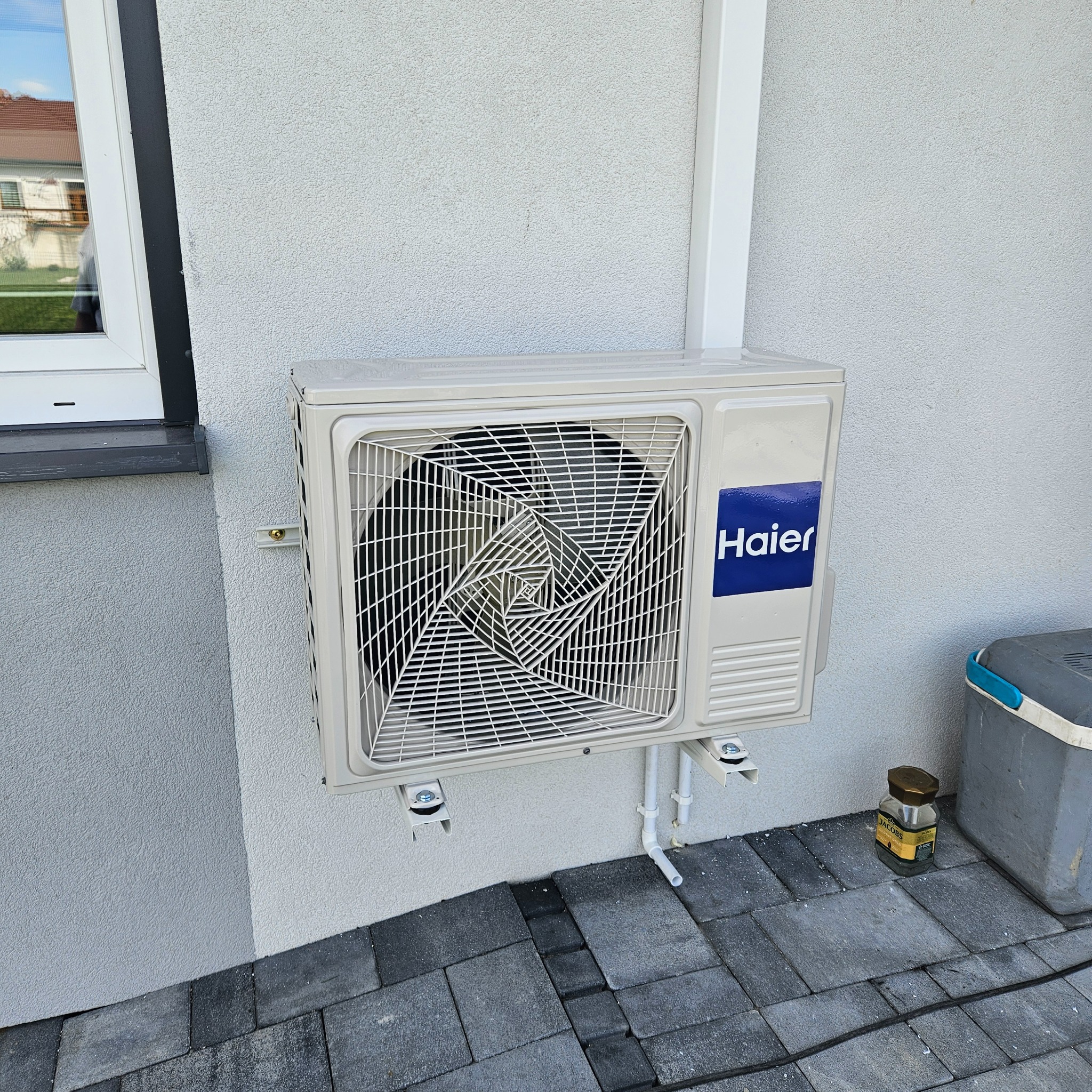 Zewnętrzna jednostka klimatyzacji Haier zamontowana na elewacji domu, widoczne rurki odprowadzające skropliny i puszka po kawie Jacobs na kostce brukowej.