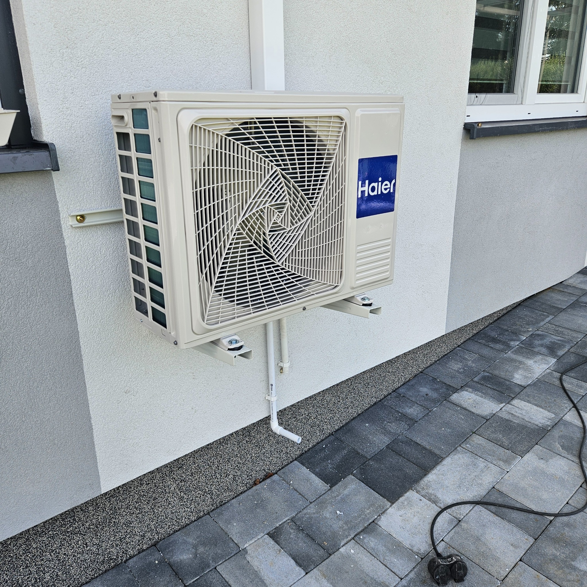Zewnętrzna jednostka klimatyzacji Haier zamontowana na elewacji domu, widoczne rury odprowadzające skropliny oraz kabel zasilający leżący na kostce brukowej.