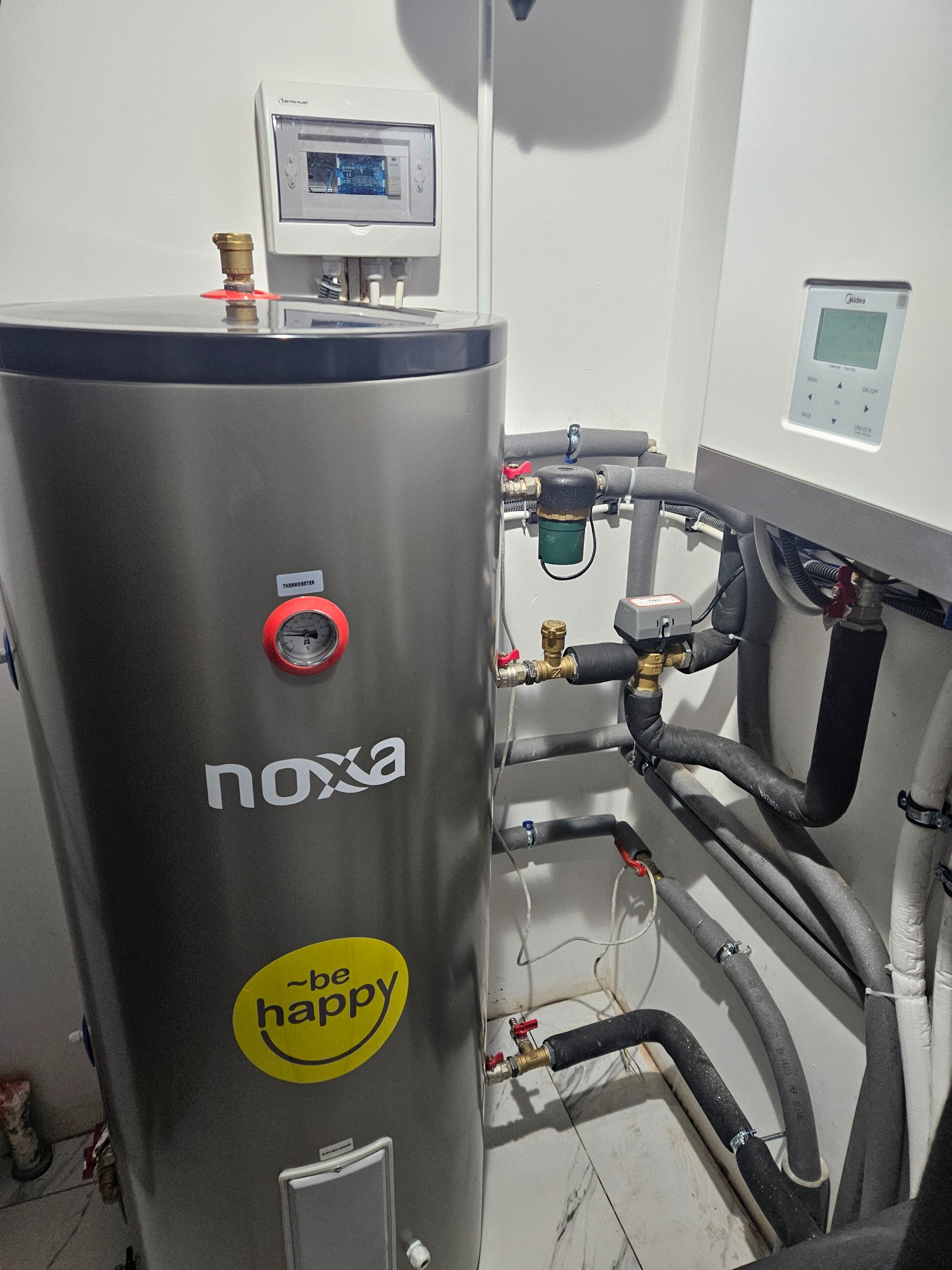 Szary zasobnik ciepłej wody użytkowej marki Noxa z naklejką 'Be Happy', podłączony do instalacji hydraulicznej z licznymi rurami, zaworami i filtrem, obok wisząca pompa ciepła Midea i panel...