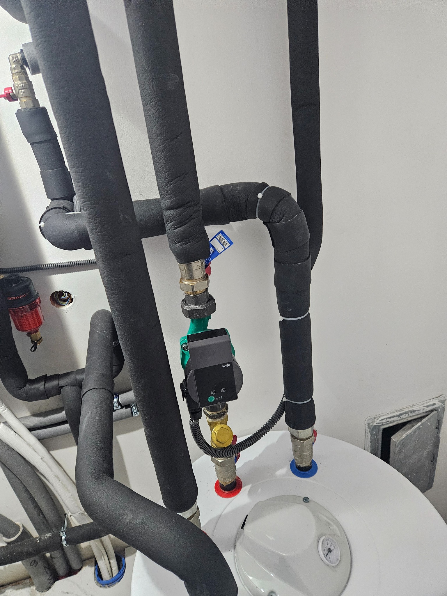 Instalacja grzewcza: rury w czarnej izolacji, zawory z kolorowymi oznaczeniami, pompa obiegowa Wilo zamontowana na białym zbiorniku, widoczne elementy instalacji hydraulicznej.