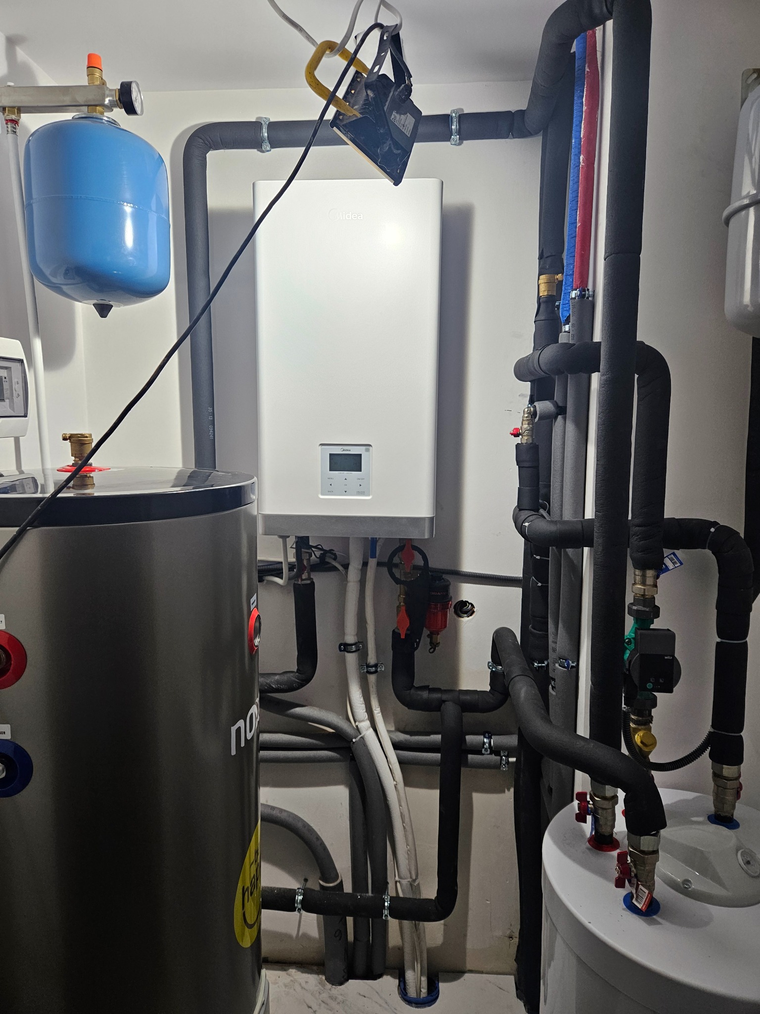 Węzeł cieplny z kotłem gazowym, zbiornikiem buforowym i zasobnikiem ciepłej wody użytkowej, rury w izolacji termicznej, manometr na niebieskim zbiorniku wyrównawczym.