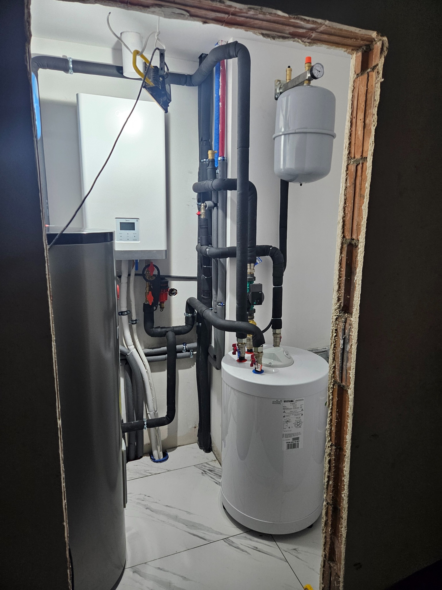 Węzeł cieplny w pomieszczeniu z wykończonymi ścianami, widoczne rury w izolacji, zbiorniki, oraz biała jednostka sterująca na ścianie, oświetlenie robocze zawieszone na rurze.