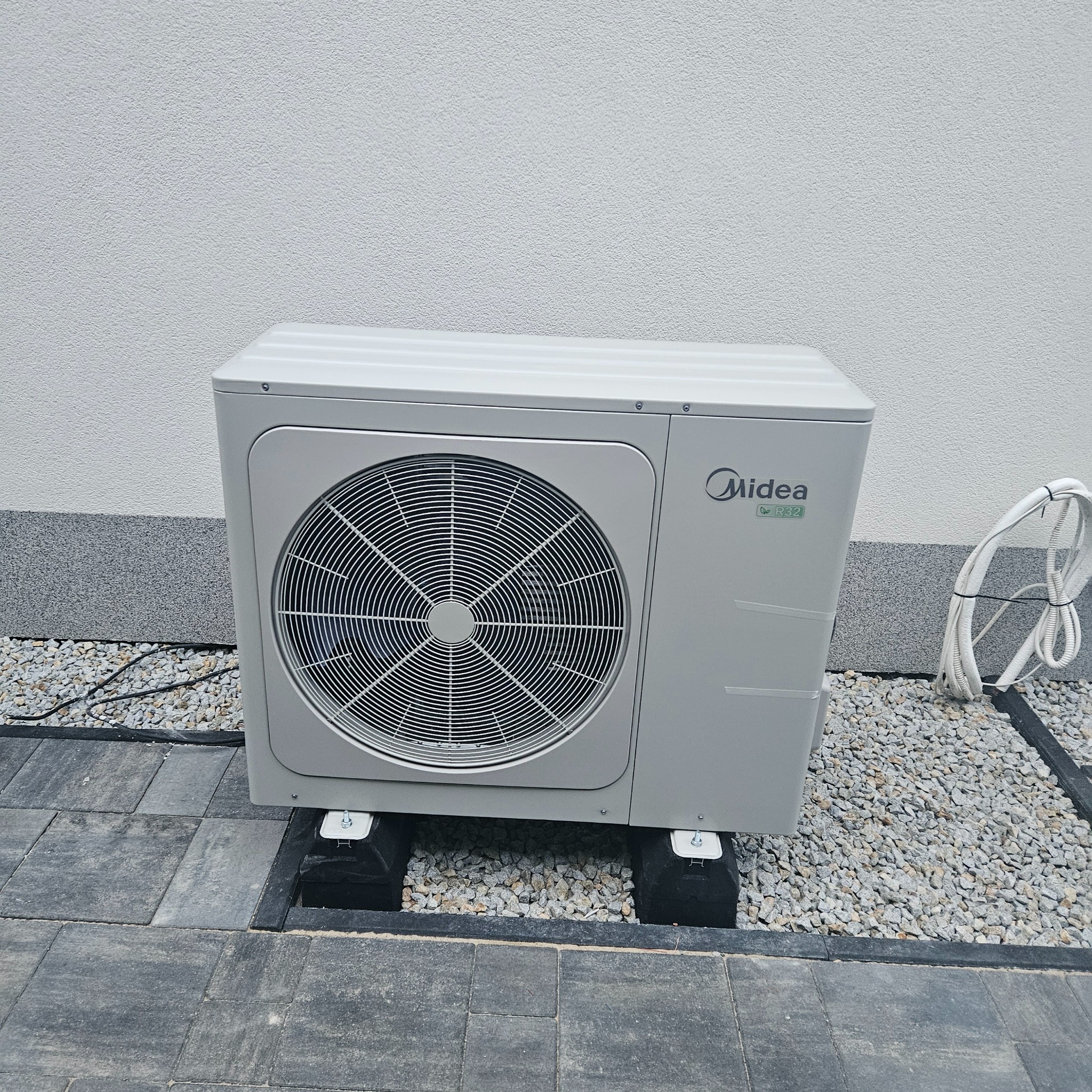 Zewnętrzna jednostka klimatyzacji Midea R32 na czarnych podkładkach, otoczona szarym żwirem i kostką brukową, przy jasnej ścianie budynku.