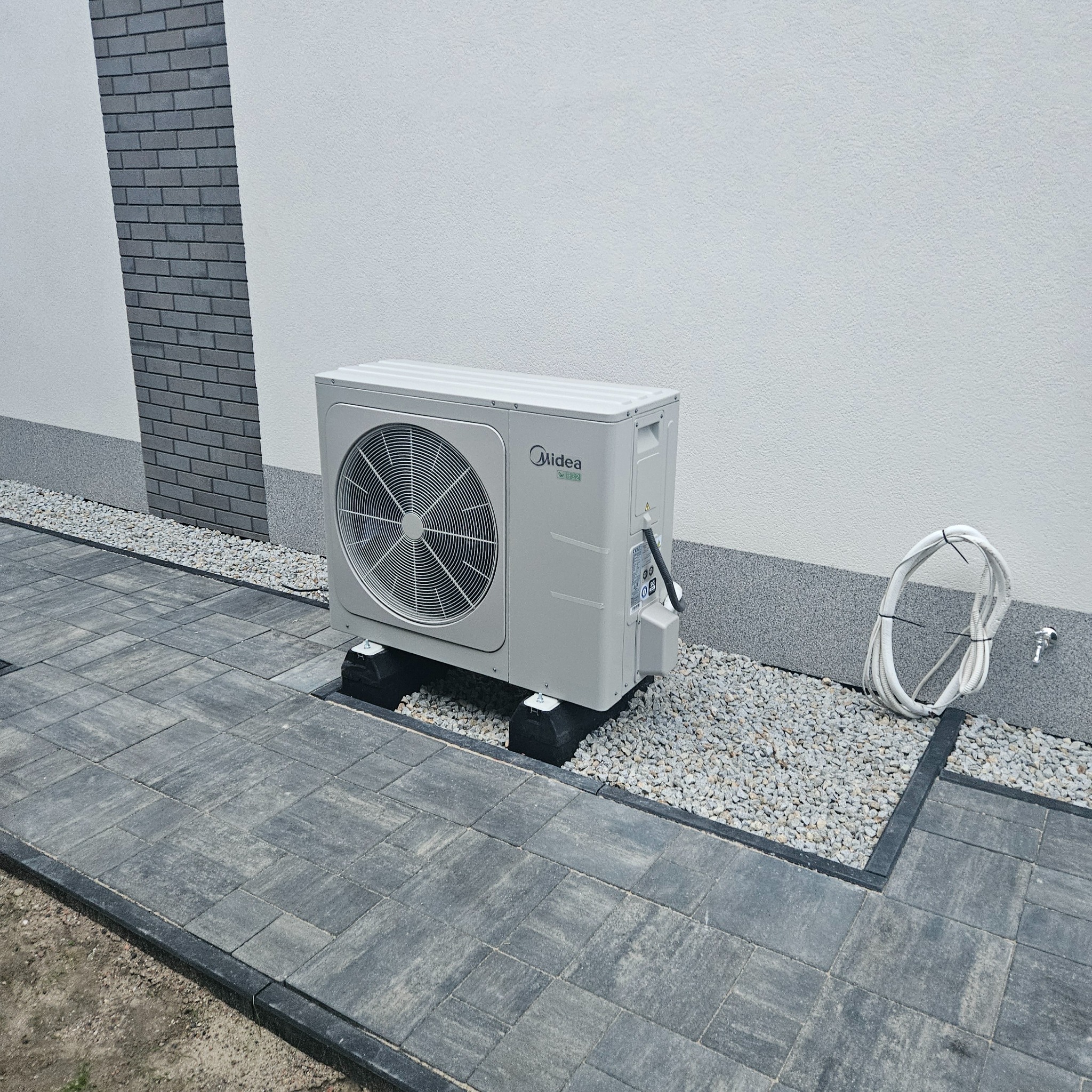 Zewnętrzna jednostka klimatyzacji Midea na czarnych podkładkach, zamontowana na szarym brukowanym chodniku z obrzeżami, obok ściany budynku z dekoracyjnym elementem z szarej cegły i zwiniętymi rurami.