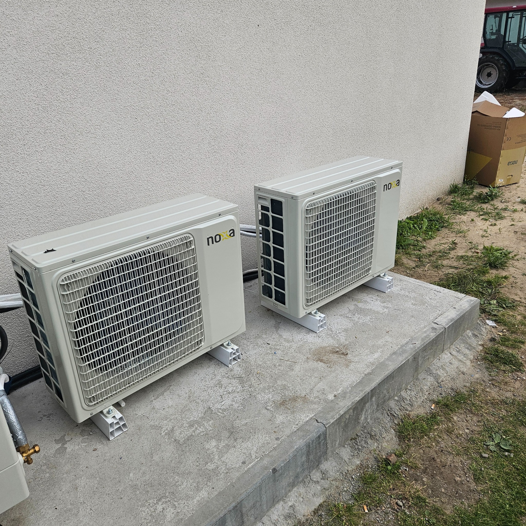 Dwa klimatyzatory marki Noxa zamontowane na betonowym fundamencie przy ścianie budynku z fakturą baranka, widoczne rury instalacyjne i kartonowe opakowanie w tle.