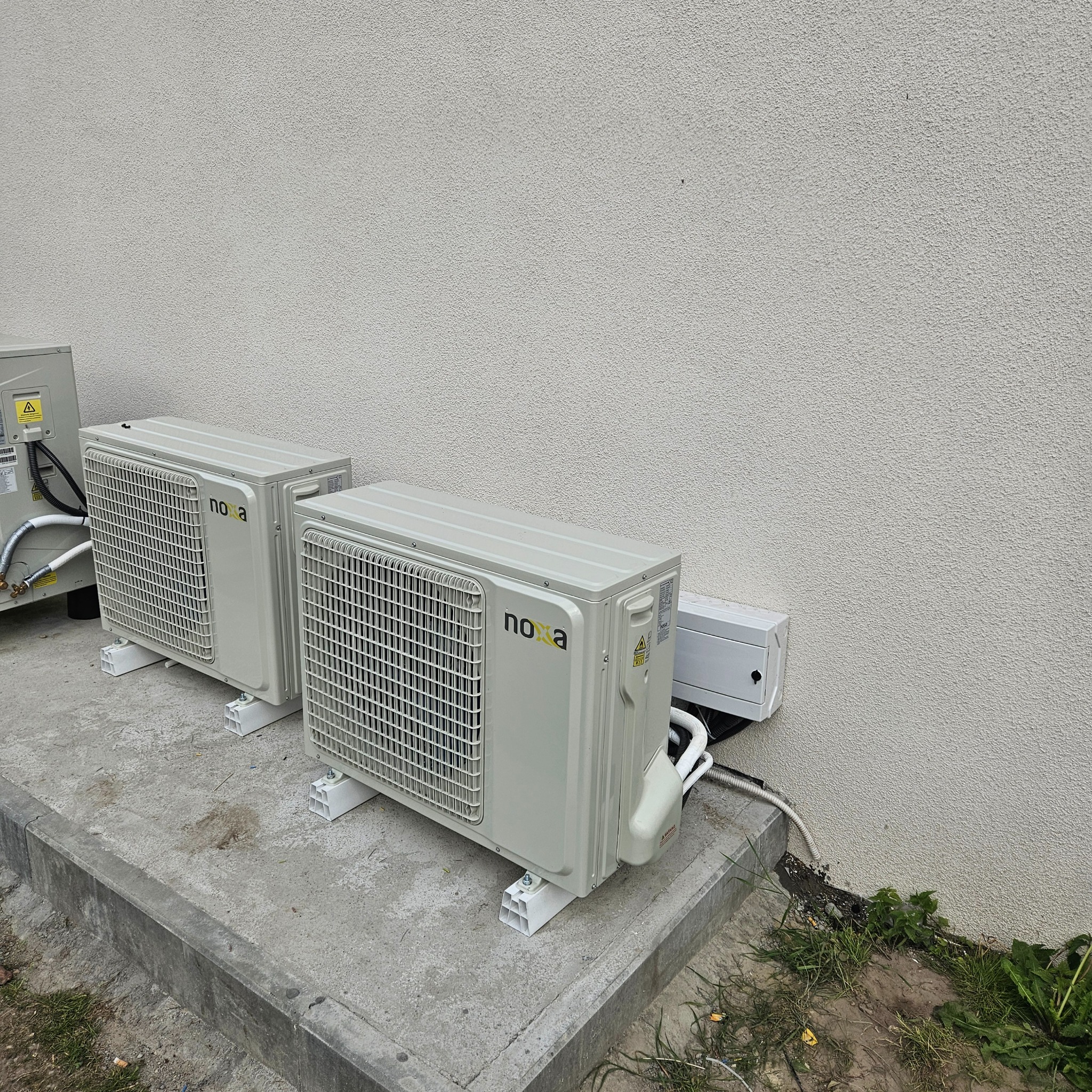 Trzy jednostki zewnętrzne klimatyzacji 'Nova' ustawione na betonowym podłożu przy jasnej, teksturowanej ścianie budynku, z widocznymi przewodami i skrzynką elektryczną.