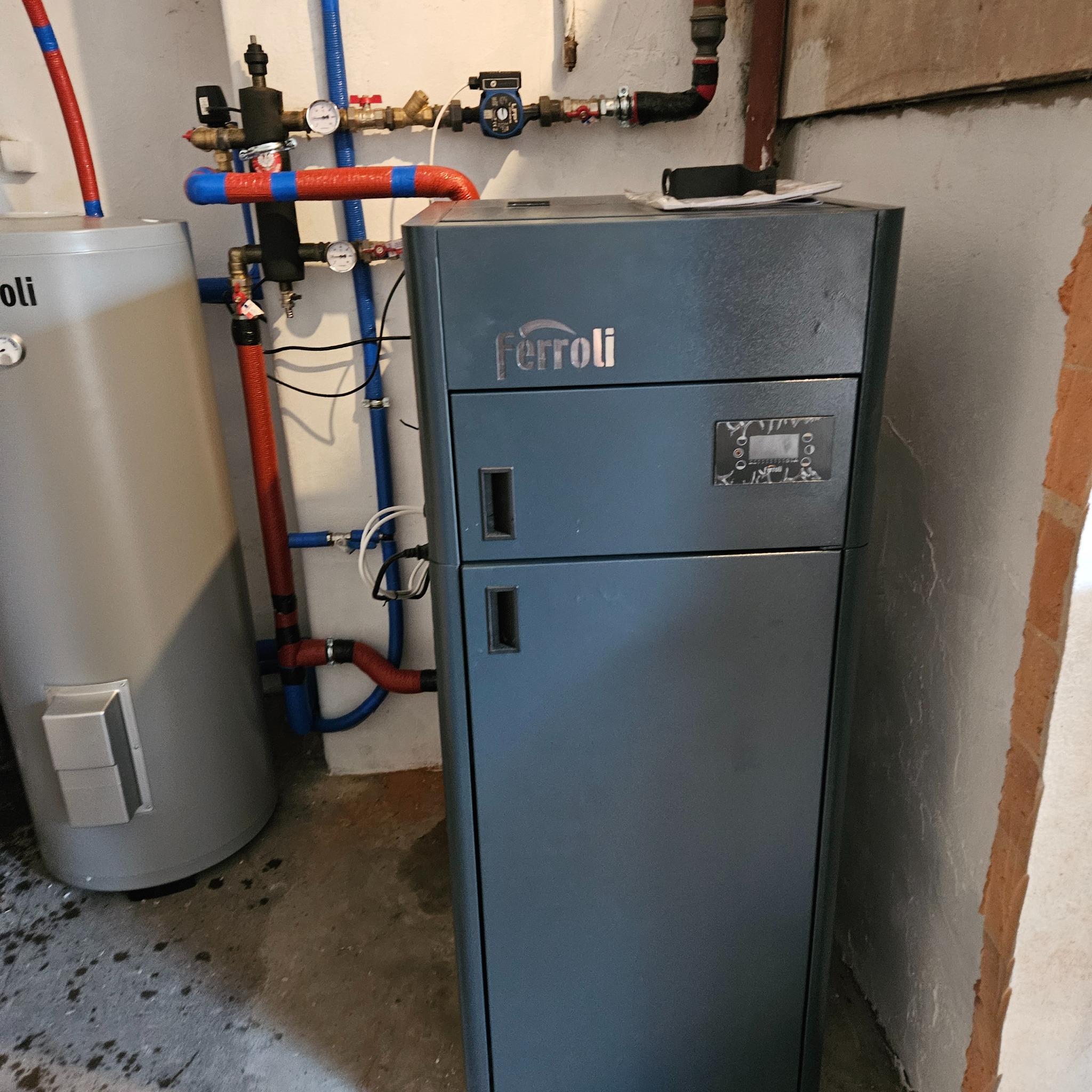 Szary piec Ferroli z widocznymi rurami instalacji grzewczej i zasobnikiem ciepłej wody, zainstalowany w pomieszczeniu z surowym wykończeniem ściany.