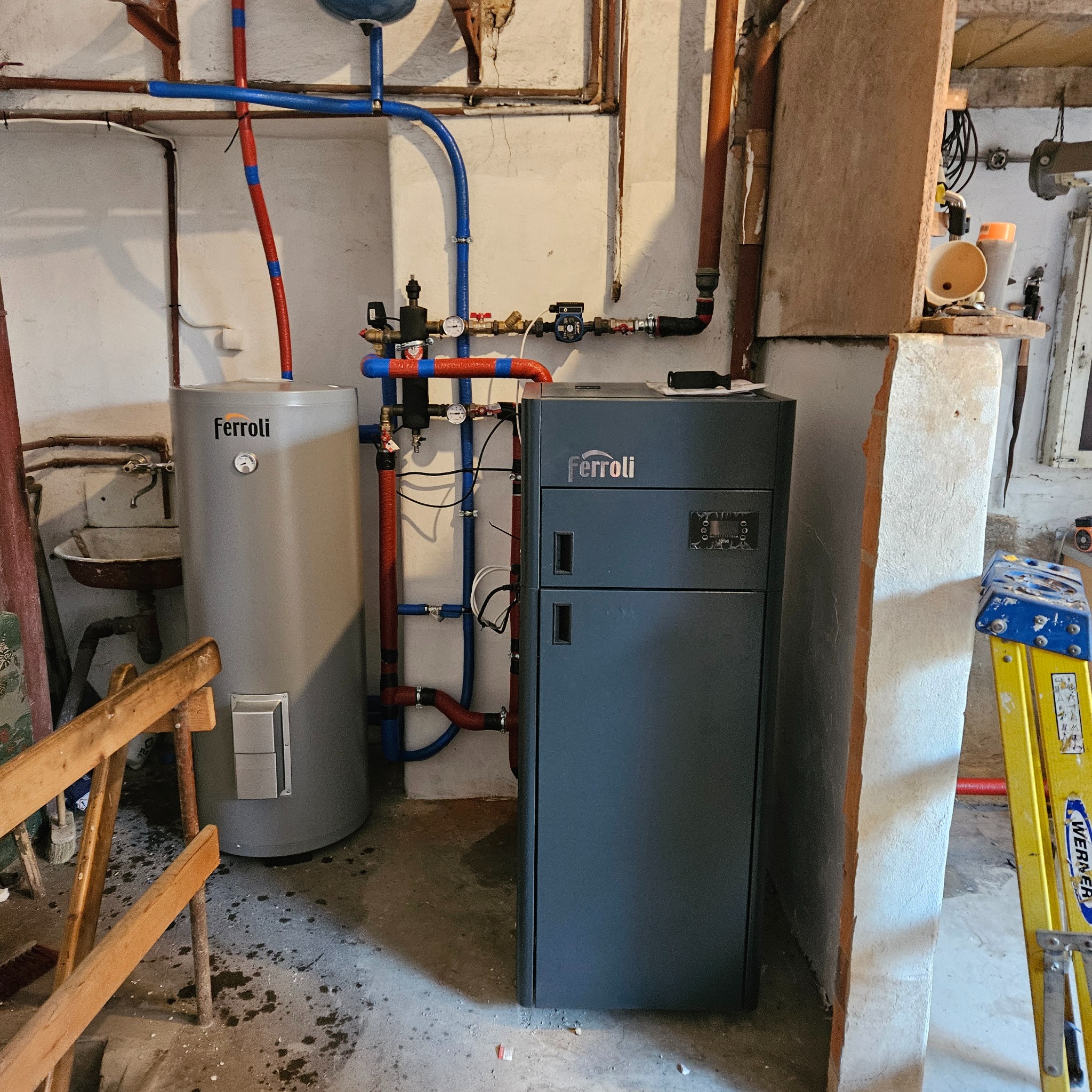 Szary piec Ferroli z podłączonym zasobnikiem ciepłej wody, widoczne rury instalacji CO i CWU w piwnicy z surowymi ścianami.