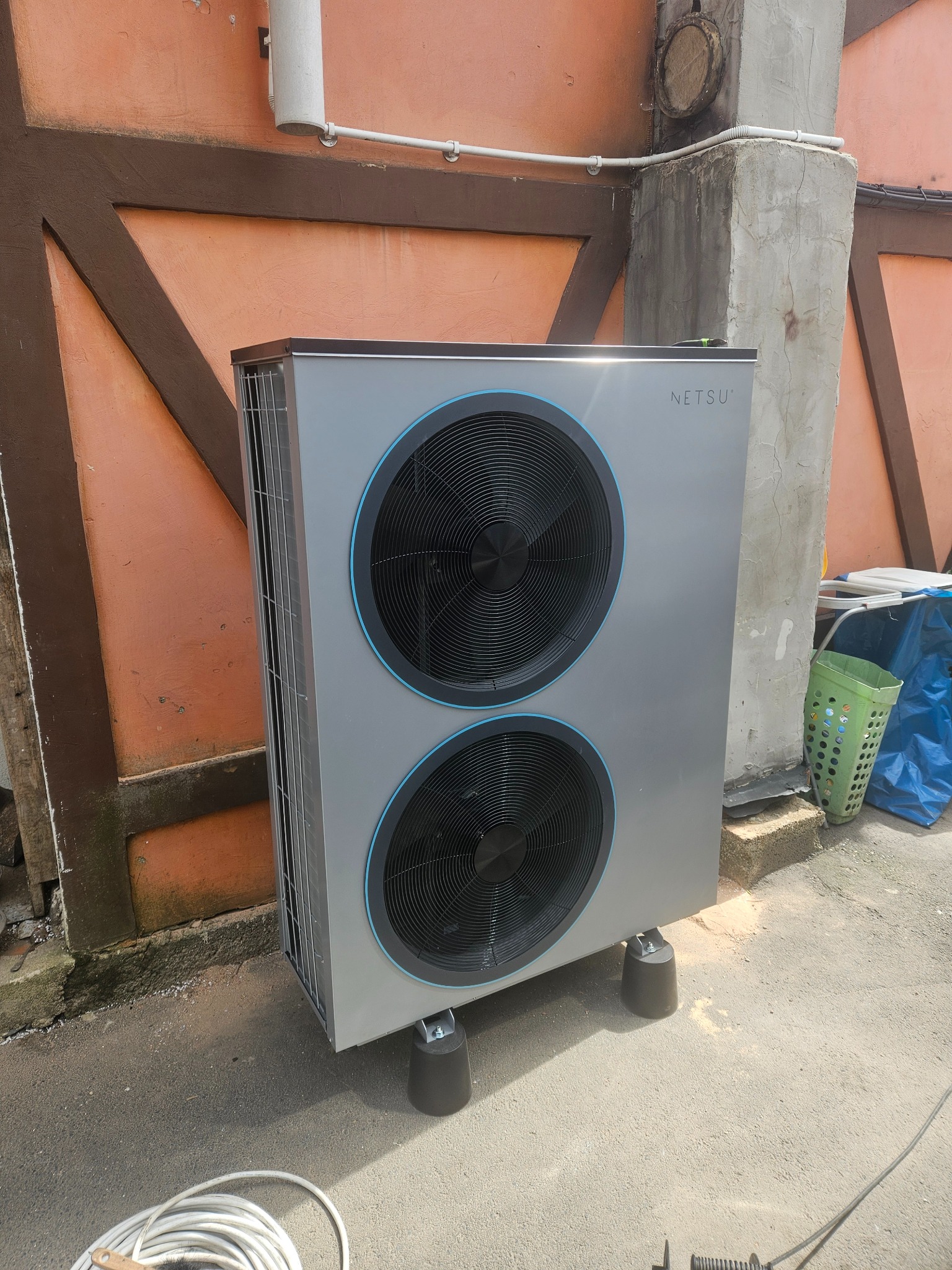 Zewnętrzna jednostka pompy ciepła NIBE ustawiona na gumowych nóżkach, widoczna na tle pomarańczowej ściany budynku z elementami dekoracyjnymi w kolorze brązowym.