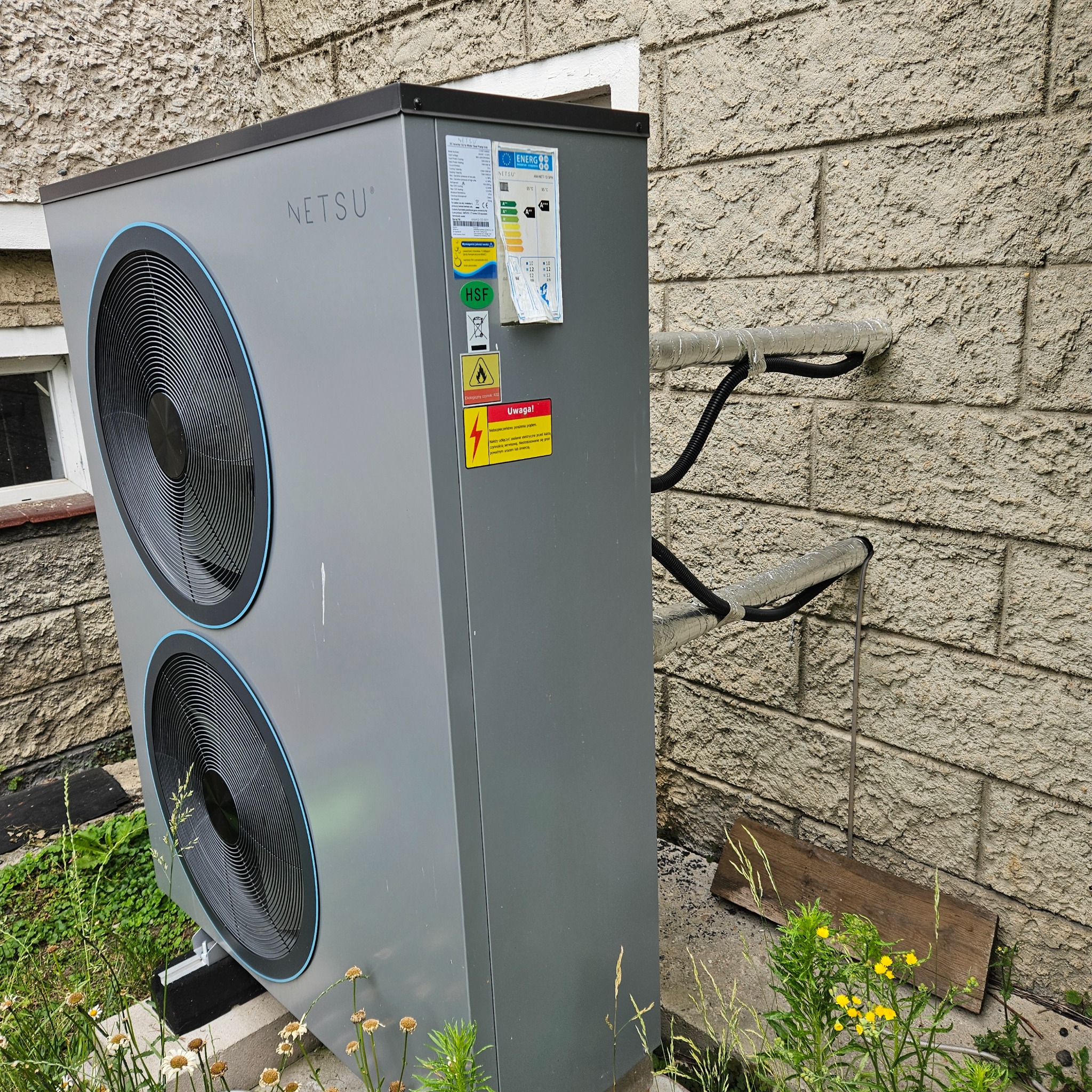 Szara pompa ciepła Netsu zamontowana na zewnątrz budynku z kremową, chropowatą elewacją, widoczne rury w izolacji termicznej wchodzące do ściany, naklejki z informacjami technicznymi...