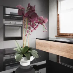 Nowoczesna kuchnia z szarymi szafkami, drewnianym blatem, oknem z roletami rzymskimi i dekoracyjną orchideą w białej donicy na błyszczącym czarnym blacie.