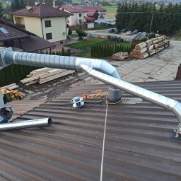 TT Inwest - Instalacja odpylania z rurami wentylacyjnymi na brązowym dachu blachodachówki, widoczna zwijarka kabla i lina zabezpieczająca.