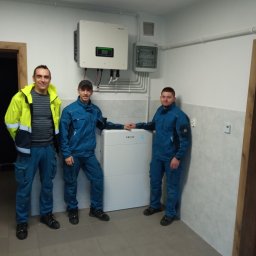 TT Inwest - Trzech mężczyzn w strojach roboczych pozuje obok białego magazynu energii z podłączonym inwerterem na ścianie w pomieszczeniu z szarą podłogą i biało-szarymi kafelkami na ścianach.