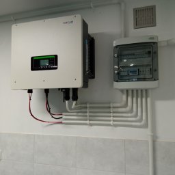 TT Inwest - Biały falownik SOFAR zamontowany na ścianie z szarą glazurą, obok rozdzielnica elektryczna w obudowie z przezroczystym okienkiem i licznymi przewodami w białych peszlach.