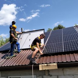 TT Inwest - Trzech monterów w uprzężach zabezpieczających instaluje panele fotowoltaiczne na brązowym blaszanym dachu domu jednorodzinnego, błękitne niebo w tle.