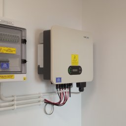 TT Inwest - Biała rozdzielnica PV AC i falownik SOFAR Solar zamontowane na ścianie, z widocznymi przewodami w peszlach i kablami podłączeniowymi, żółte naklejki ostrzegawcze o napięciu.