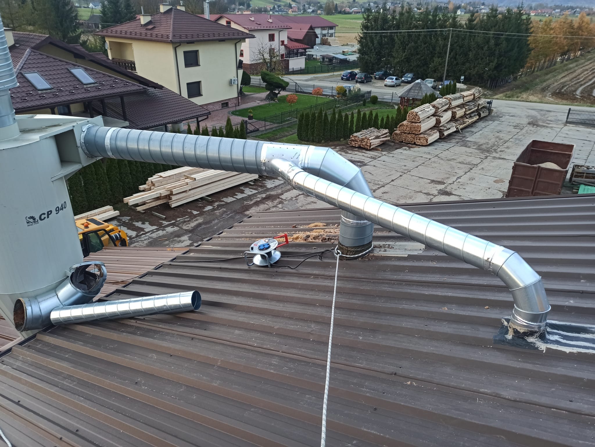 Instalacja odpylania z rurami wentylacyjnymi na brązowym dachu blachodachówki, widoczna zwijarka kabla i lina zabezpieczająca.