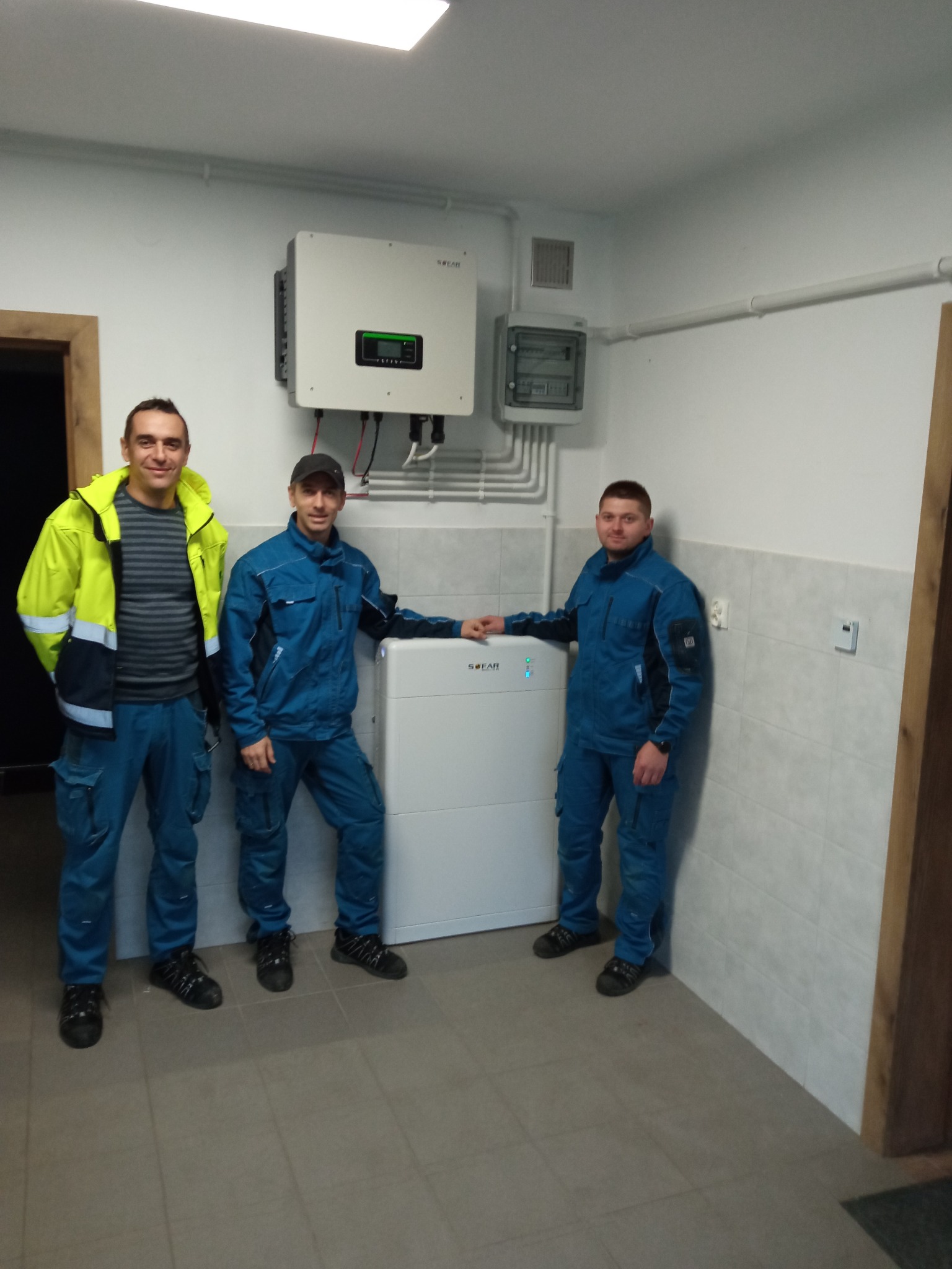Trzech mężczyzn w strojach roboczych pozuje obok białego magazynu energii z podłączonym inwerterem na ścianie w pomieszczeniu z szarą podłogą i biało-szarymi kafelkami na ścianach.