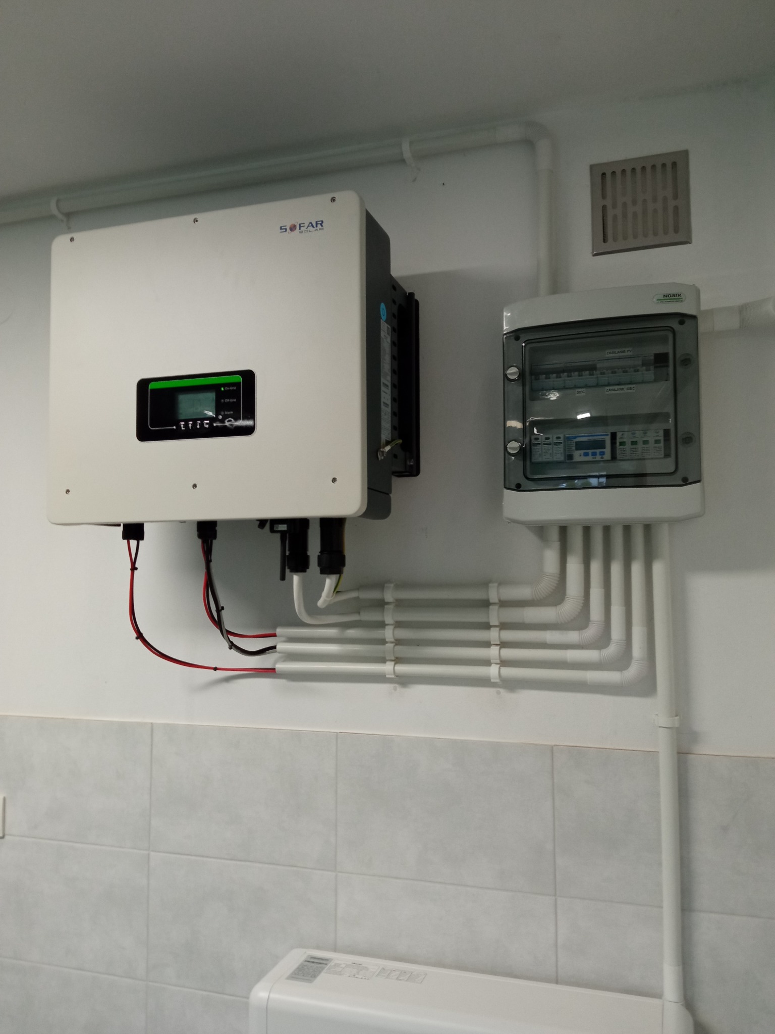 Biały falownik SOFAR zamontowany na ścianie z szarą glazurą, obok rozdzielnica elektryczna w obudowie z przezroczystym okienkiem i licznymi przewodami w białych peszlach.