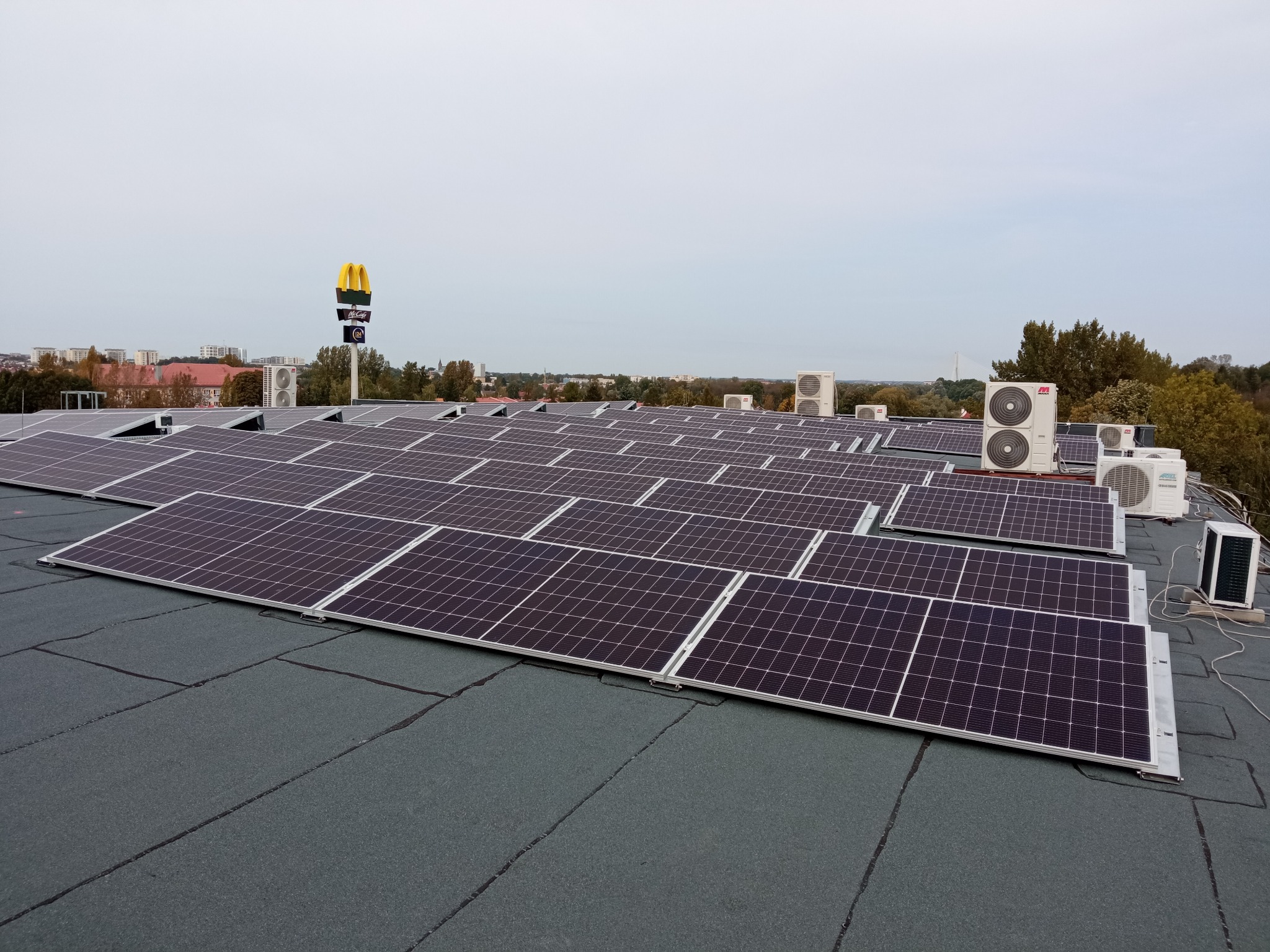 Rząd paneli słonecznych na płaskim dachu budynku z widocznymi jednostkami klimatyzacyjnymi i logo McDonald's w tle.