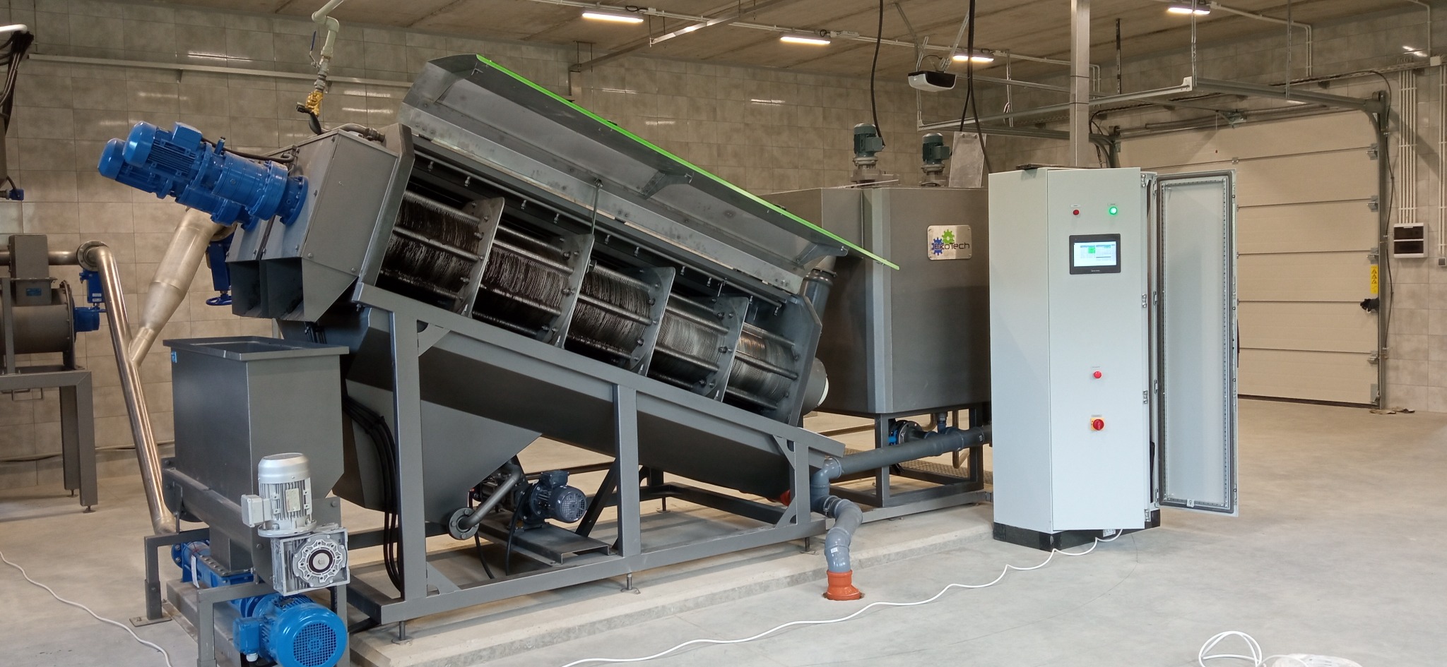 Szara maszyna przemysłowa z niebieskimi silnikami i panelem sterowania z otwartymi drzwiami, stojąca w hali z szarymi płytkami i bramą garażową w tle.