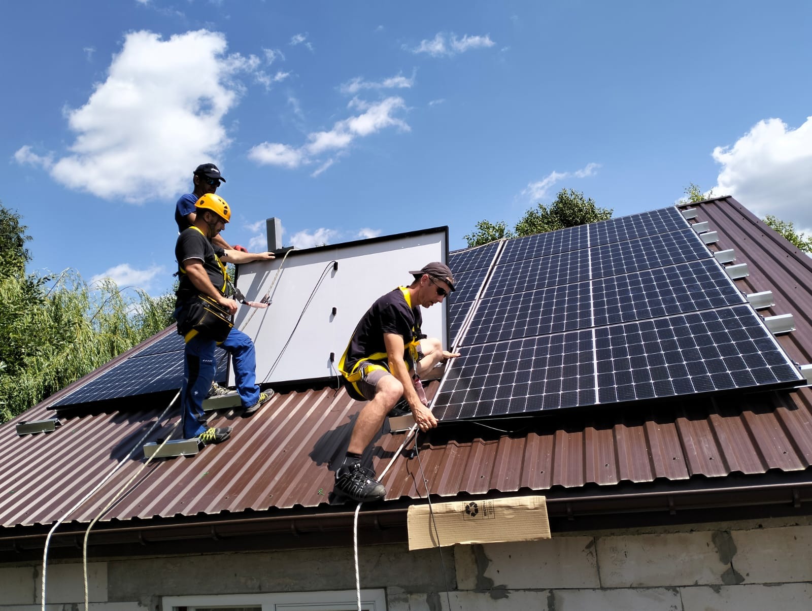 Trzech monterów w uprzężach zabezpieczających instaluje panele fotowoltaiczne na brązowym blaszanym dachu domu jednorodzinnego, błękitne niebo w tle.