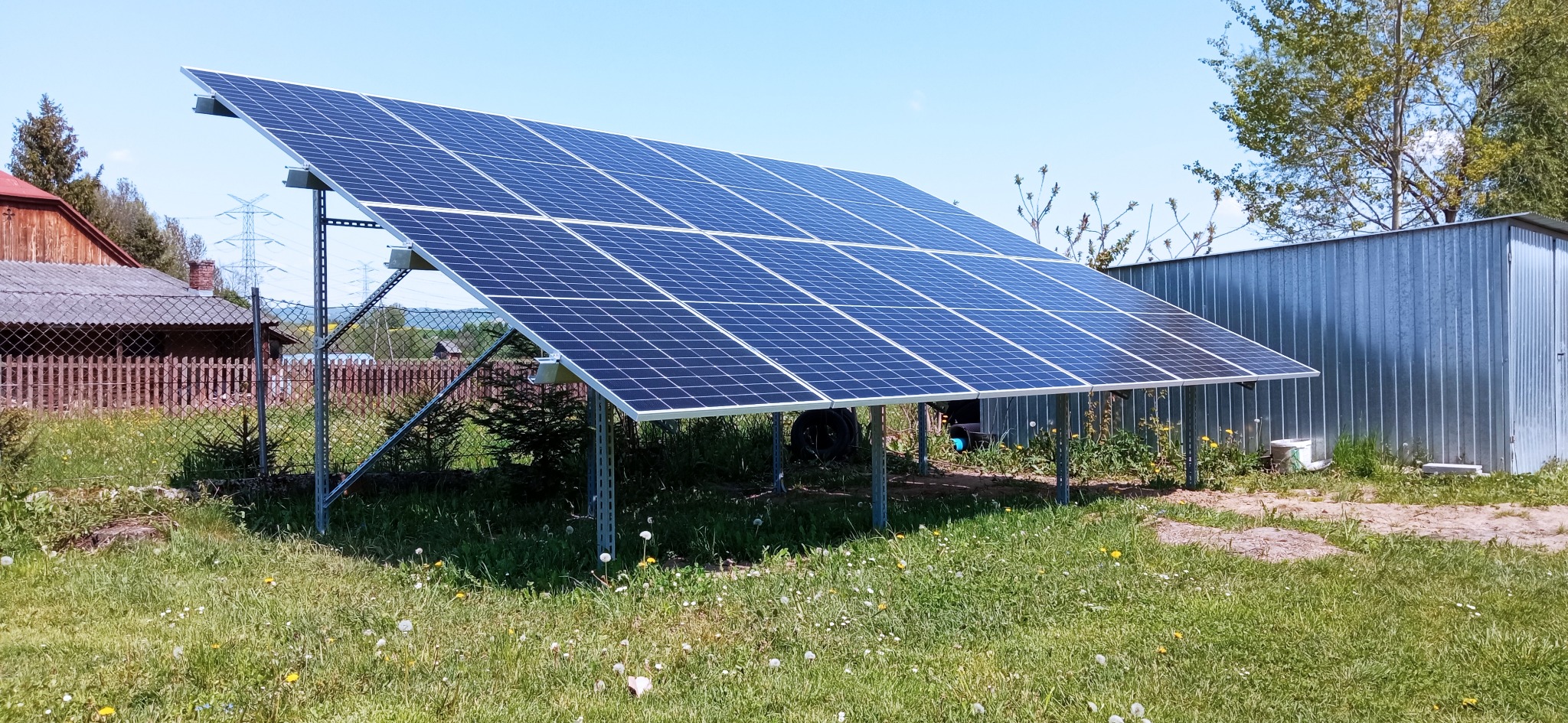 Panel fotowoltaiczny na metalowej konstrukcji, zainstalowany na trawiastym terenie obok blaszanej szopy, z widocznymi drzewami i liniami wysokiego napięcia w tle.
