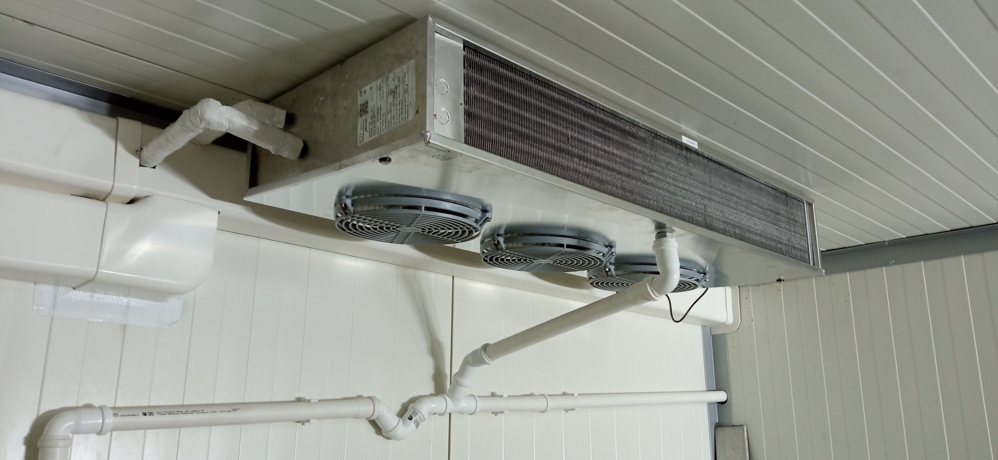 Chłodnica przemysłowa z trzema wentylatorami zamontowana pod sufitem chłodni, widoczne białe rury instalacji chłodniczej.