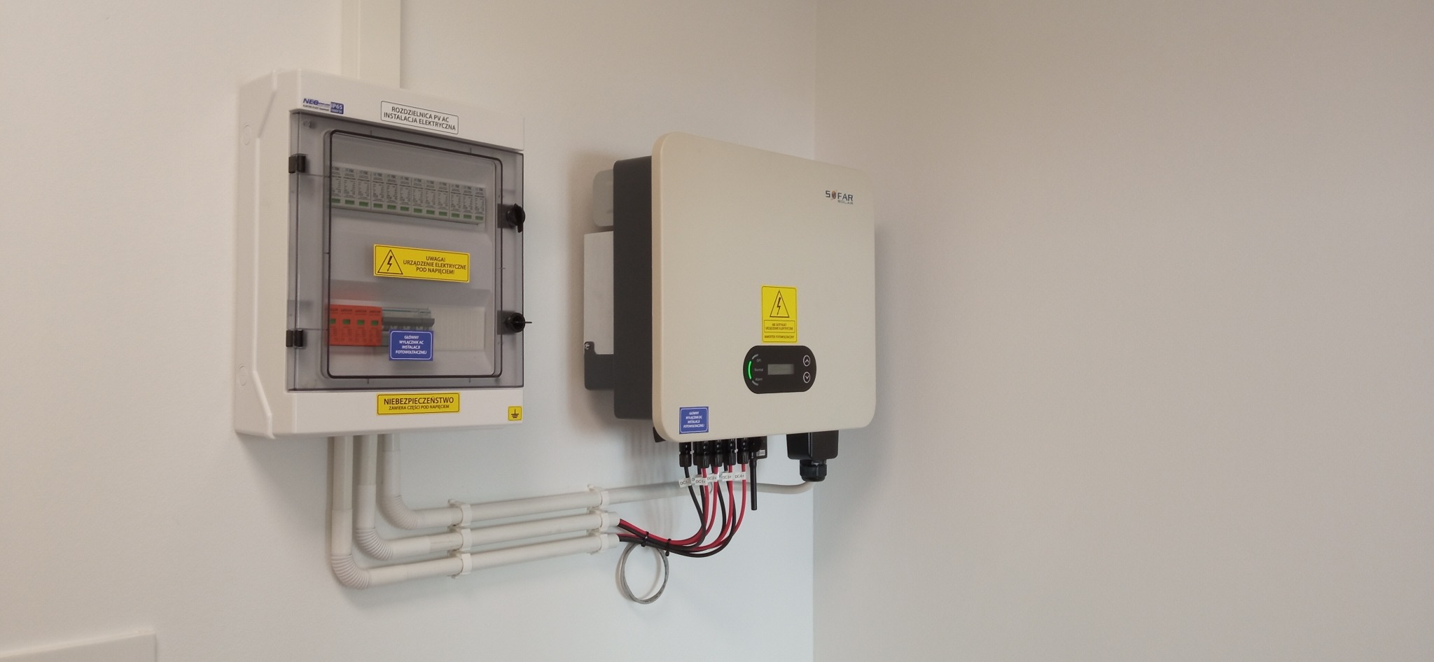 Biała rozdzielnica PV AC i falownik SOFAR Solar zamontowane na ścianie, z widocznymi przewodami w peszlach i kablami podłączeniowymi, żółte naklejki ostrzegawcze o napięciu.