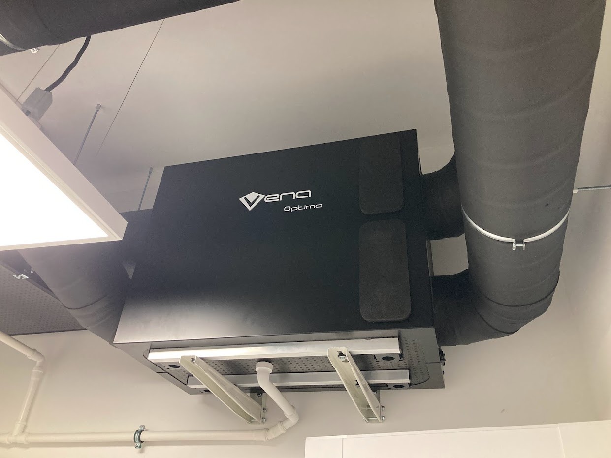 Czarna centrala wentylacyjna Vena Optima zamontowana pod sufitem, widoczne izolowane rury wentylacyjne i elementy mocujące.