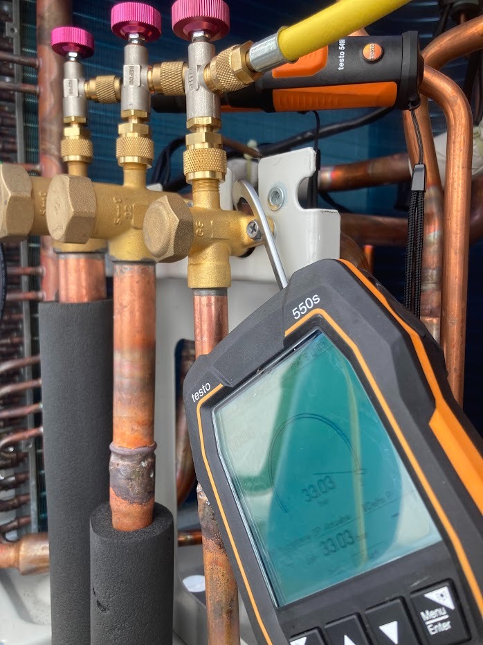 Pomiar parametrów instalacji chłodniczej za pomocą elektronicznego manometru Testo 550s, widoczne zawory z różowymi pokrętłami i miedziane rury.