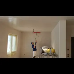 Pracownik w niebieskiej koszulce i białych spodniach, stojąc na podwyższeniu, wyrównuje świeżo nałożoną masę szpachlową na suficie za pomocą długiego narzędzia z czerwoną końcówką. Okno...