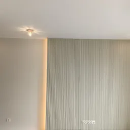 Ściana z dekoracyjnymi panelami pionowymi w kolorze beżowym, oświetlona listwą LED po lewej stronie, z widocznymi gniazdkami elektrycznymi i dwoma lampami sufitowymi.