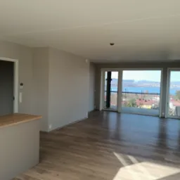 Jasne, przestronne wnętrze z drewnianą podłogą, świeżo pomalowanymi ścianami w odcieniach szarości i dużym oknem balkonowym z widokiem na miasto i jezioro.