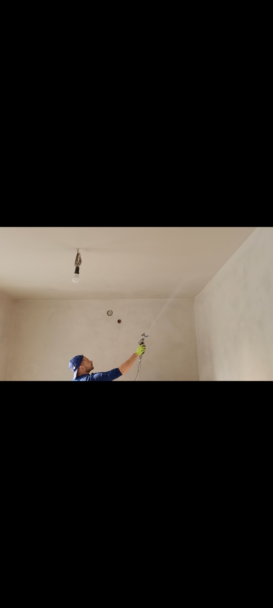 Pracownik w niebieskiej czapce, z pistoletem natryskowym w dłoni, aplikuje gładź na ścianę w pomieszczeniu z wiszącą żarówką.