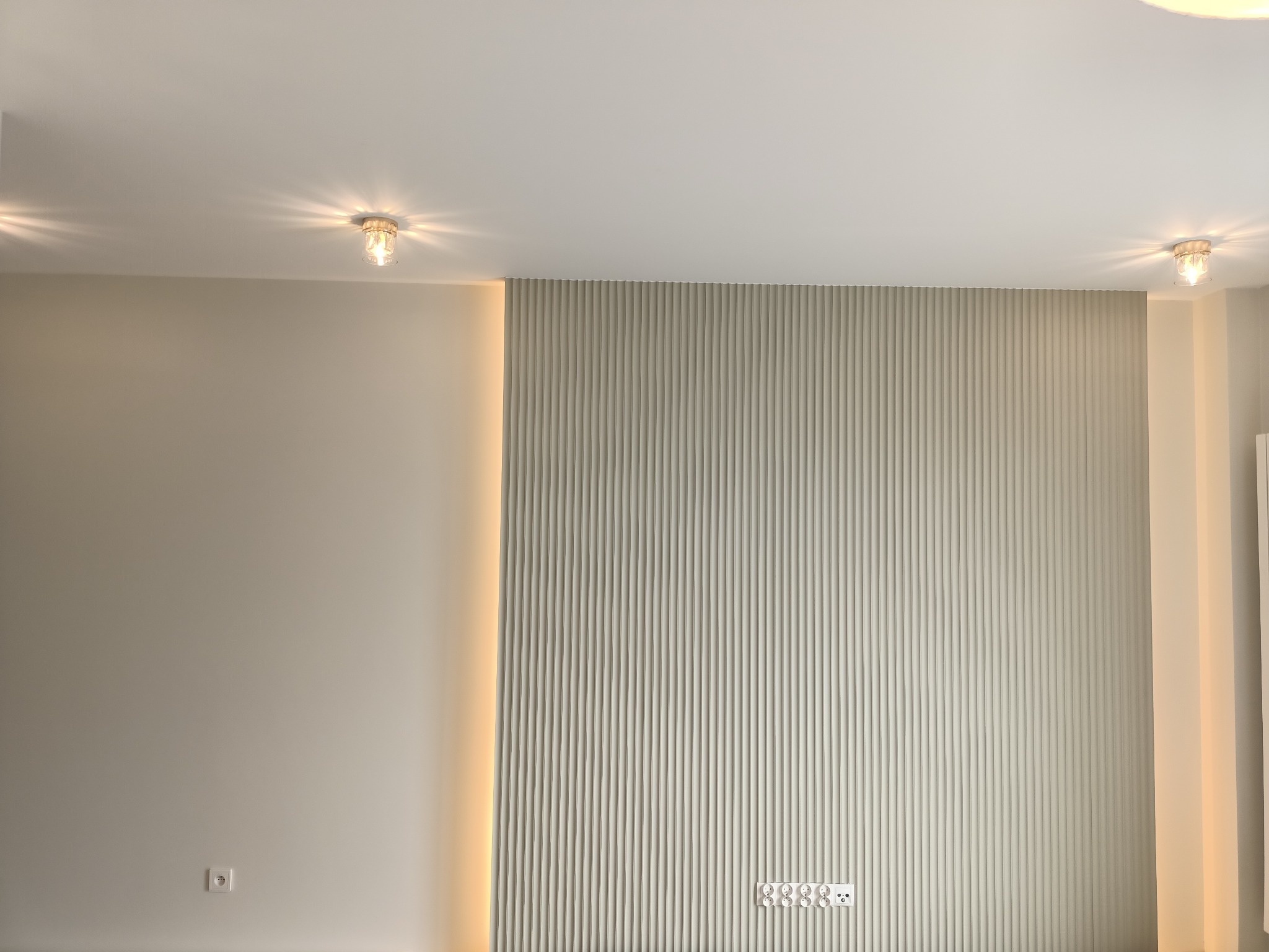 Ściana z dekoracyjnymi panelami pionowymi w kolorze beżowym, oświetlona listwą LED po lewej stronie, z widocznymi gniazdkami elektrycznymi i dwoma lampami sufitowymi.