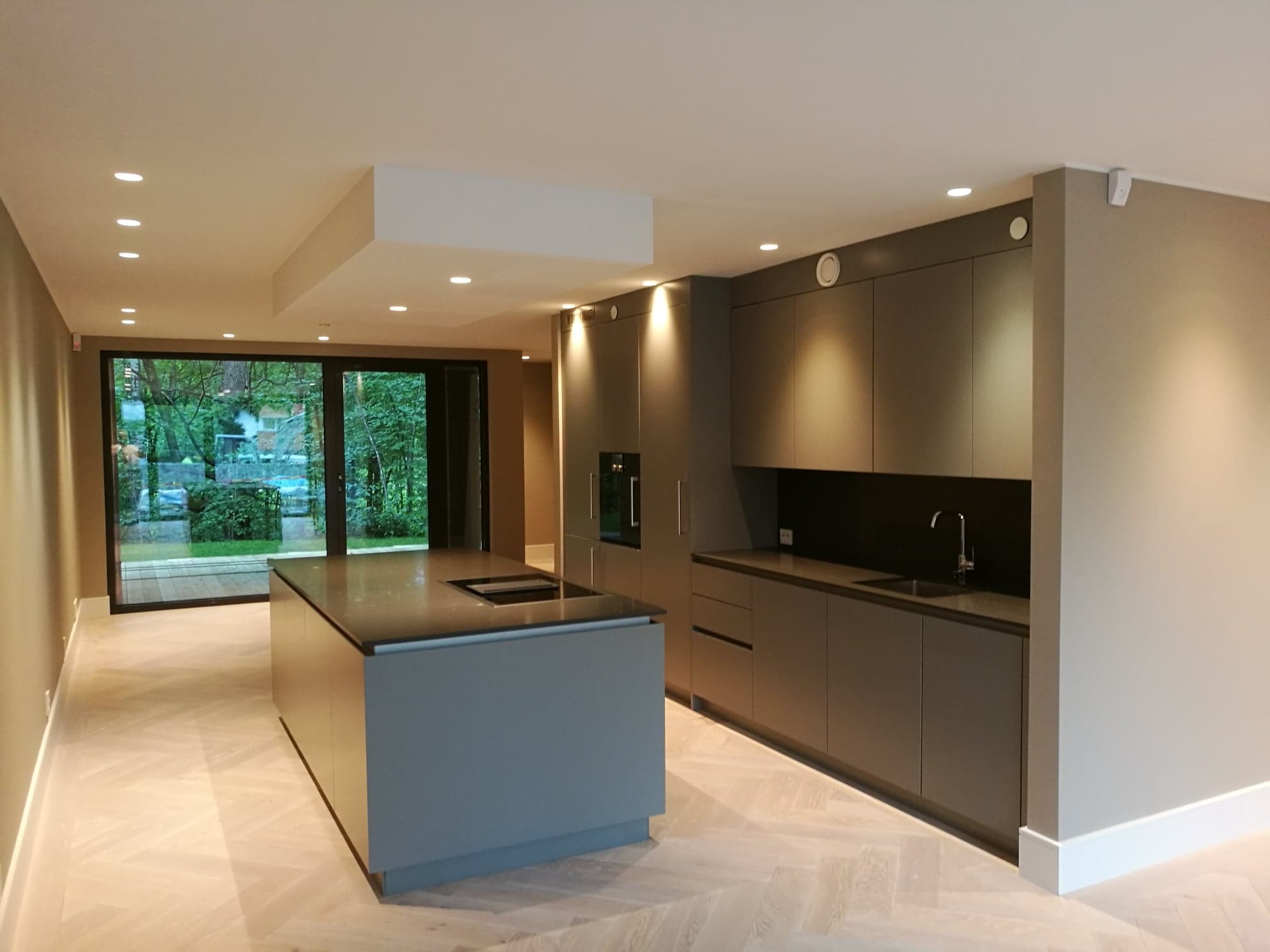 Nowoczesna kuchnia z szarymi szafkami, wyspą kuchenną z czarnym blatem i parkietem ułożonym w jodełkę, oświetlona punktowymi lampami sufitowymi.